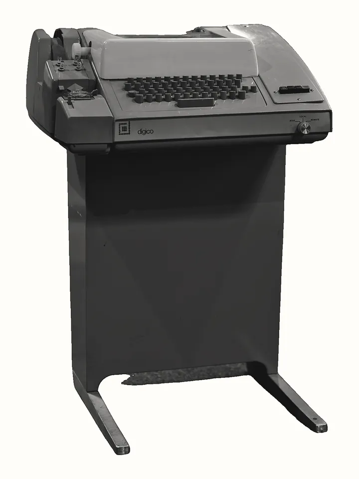Một máy teletype đứng trên chân sắt: bàn phím, khoang in giấy cuộn phía trên, bộ đọc băng giấy bên trái.